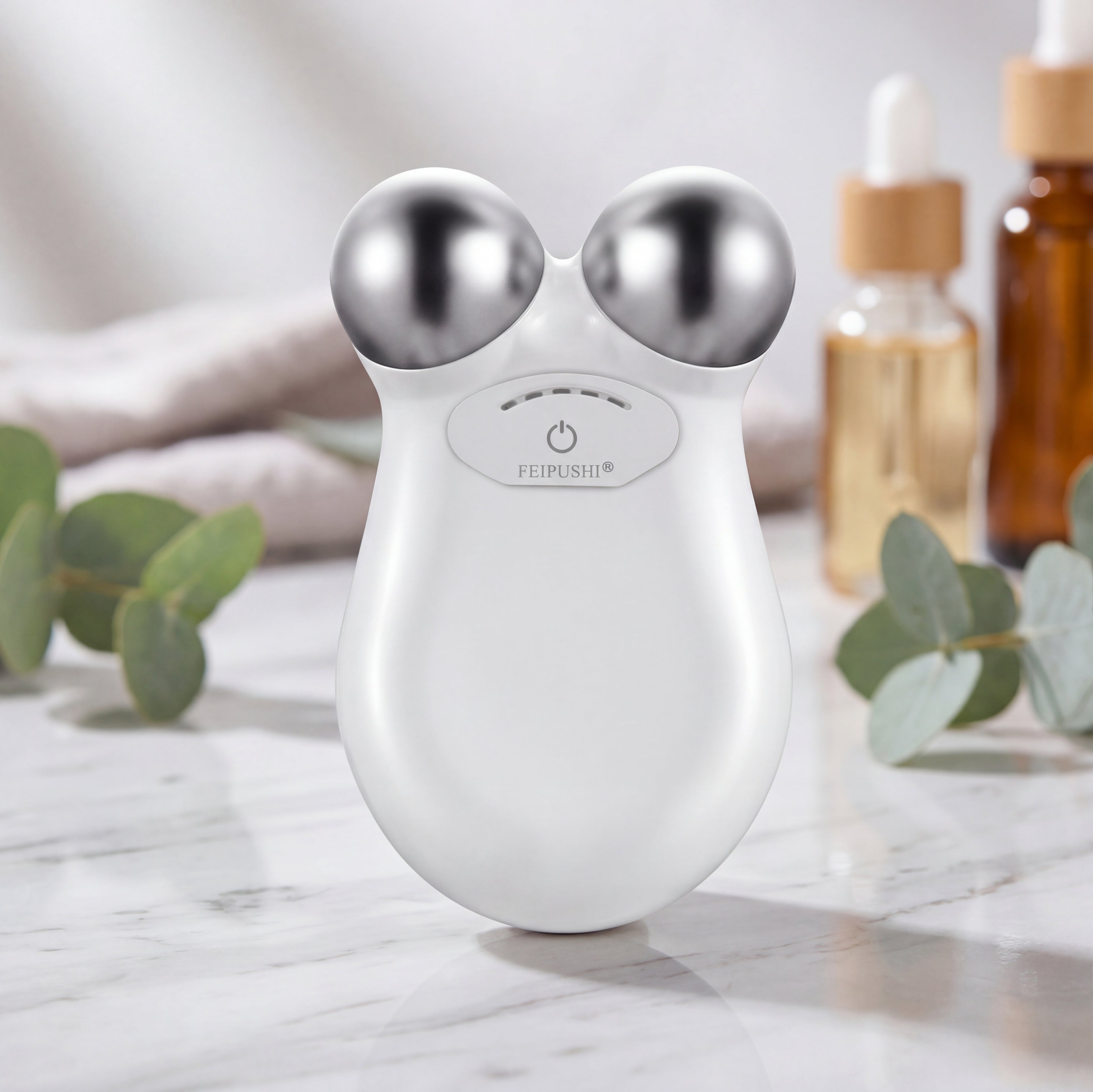 White facial massager with silver knobs on a marble surface with eucalyptus leaves and bottles in the background.